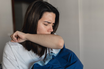 Young ill woman infected by the coronavirus is coughing into her upper sleeve while covering with a blue quilt in her home
