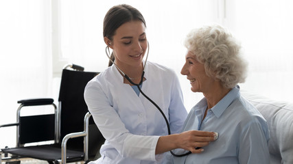 Positive female caregiver visit smiling senior disabled grandmother check heart rate, young woman...