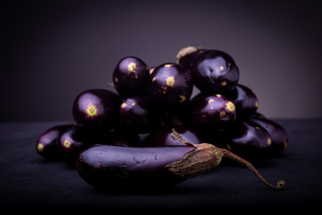 Purple baby eggplant on a black surface with a pile of aubergine vegetables out of focus in the background with one in front. Studio low key food image.