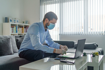Young adult one man caucasian wearing gloves on hands and protective mask on face while working from home sitting by the table laptop preventing virus spread in epidemic quarantine health prevention