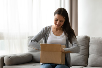 Happy young woman sit on couch in living room unpack cardboard box buying goods on Internet,...