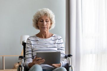 Mature disabled grandmother sit in wheelchair browsing Internet on tablet in hospital or home, smart senior old lady handicapped in invalid carriage surfing watching video on Pad, technology concept