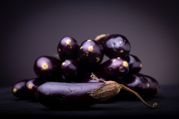 Purple baby eggplant on a black surface with a pile of aubergine vegetables out of focus in the background with one in front. Studio low key food image.