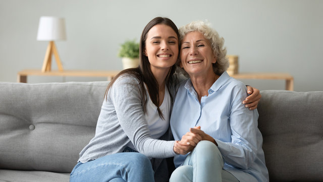 Family portrait of happy mature mom hug cuddle with grown-up adult daughter sit together on couch in living room, smiling elderly mother and millennial girl child embrace posing for picture at home