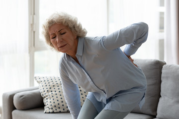 Hurt senior woman stand from couch touch lower spine suffer from sudden muscle spasm or strain,...