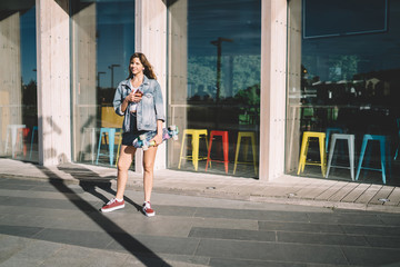 Joyful teenager with skateboard listening to music while using smartphone on street in sunny day