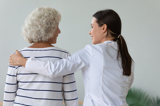 Back view of supportive young woman doctor hug comfort sick senior old lady in retirement house, female nurse or caregiver support take care of ill mature grandmother, elderly healthcare concept