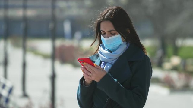 Provident brunette girl in a protective mask touching her smartphone on a quay 