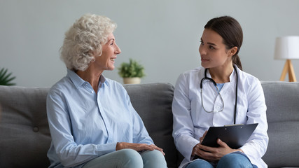 Young female doctor visit elderly woman pensioner at home give help talk sitting on couch,...