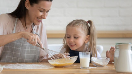 Smiling young mum stirring eggs flour milk with whisk teaching small preschooler daughter cooking...