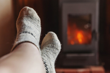 Feet legs in winter clothes wool socks at fireplace background. Woman sitting at home on winter or autumn evening relaxing and warming up. Winter and cold weather concept. Hygge Christmas eve.