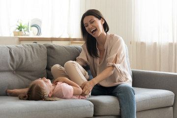 Overjoyed young mother play with cute little daughter tickle her enjoying leisure time in living room, smiling mom or nanny have fun sitting on couch with small girl child engaged in funny activity