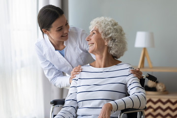Happy female nurse or caregiver talk support positive handicapped old lady patient sitting in...