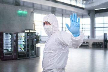 epidemiologist in a protective suit shows a prohibition gesture against the waiting room at the airport or train station - obrazy, fototapety, plakaty