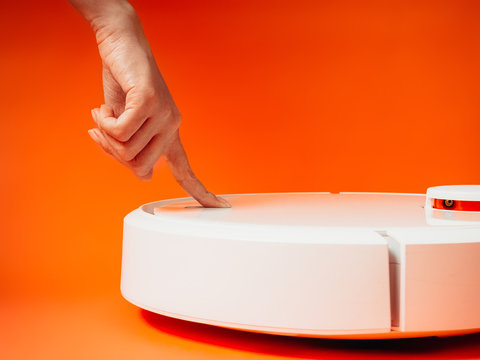 Girl presses a hand the power button on a robot vacuum cleaner on an orange background.