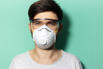 Studio portrait of young guy wearing medical face mask and protective goggles against coronavirus isolated on background of aqua menthe color.