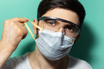 Studio close-up portrait of young guy put on medical flu mask on face, wearing protective goggles against coronavirus, isolated on background of aqua menthe color.