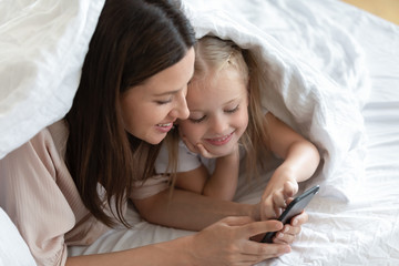 Smiling little daughter relax enjoying time with young mom playing on smartphone in bedroom...