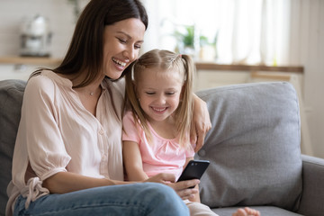 Excited young mother and little preschooler daughter sit relax on couch watching funny video on cellphone, smiling happy mom and girl child enjoy leisure time using smartphone at home together