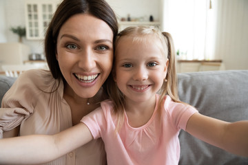Smiling young mother and small preschooler daughter look at camera making selfie at home together, happy funny mom and little girl child have fun laugh take photo posing for self-portrait picture