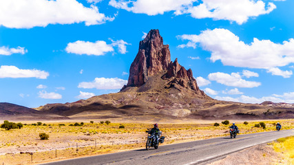 Touring Bike Riders passing the rugged peaks of El Capitan and Agathla Peak towering over the desert landscape south of Monument Valley along Highway US Route 163 in northern Arizona, United States