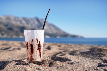 Empty glass with straw after refreshing milk cocktail with vanilla ice cream and chocolate topping is on sand on the beach on hot sunny day. Blurred sea, clear blue sky and mountain in the background