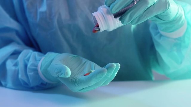 Coronavirus pandemic. Drug dosage. Doctor hands in protective gloves scattering pills from tub.