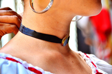 The hands of an old woman is putting a blue collar shaped like a drop or a tear in the nek of a young white lady with soft sking dressed in white dress and big earrings.