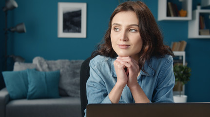 Portrait of musing young woman with her head rested on fists sitting in front of computer at home