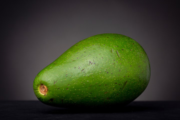 Large pear-shaped vibrant green avocado laying on a black surface contrasted against a grey background showing in detail its outer peel skin