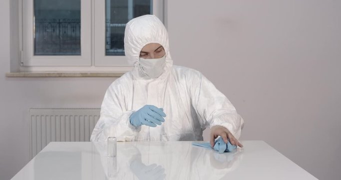 Portrait of young male person stitting behind white dining desk in kitchen in protective suit removing gloves and desinfecting his hands with an antiseptic during pandemic coronavirus quarantine time