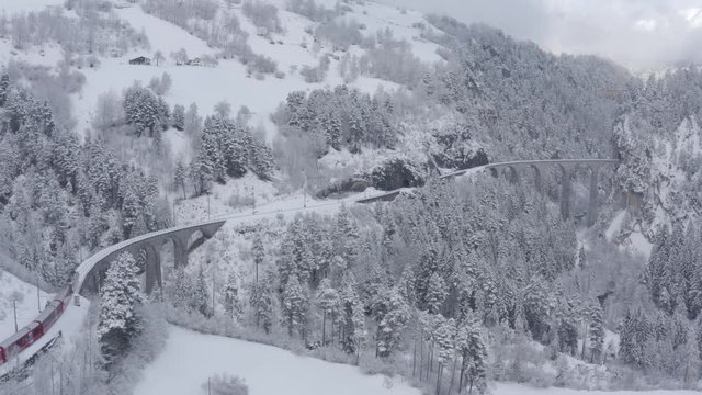 Aerial video of famous train of red color Glacier Express at winter, The train moves to the Landwasser Viaduct and tunnel, landmark of Switzerland, snowing, drone flies forward