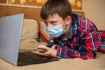 teenage boy in protective medical mask coughs in fist. child remotely does lessons lying on bed near laptop. Online shopping, e-learning, remote studying on internet concept.