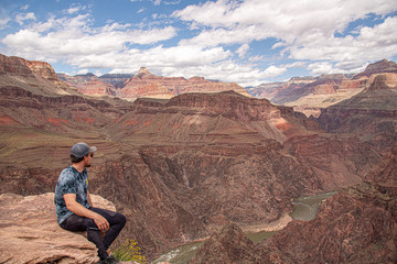 Man sitting on the edge of the Grand Canyon in Arizona during summer time with amazing Colorado River in background below. Summer time, road trip travel area in United States of America. 