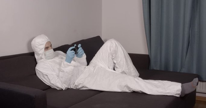 Portrait of young male person relaxing and resting on a black living room couch in protective suit playing video games on a gaming console during pandemic coronavirus quarantine time 4K