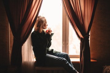 Young thoughtful woman drinking tea and looking through window while sitting on windowsill at home