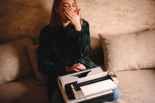 Young happy female writer laughing while using typewriter sitting on sofa at home