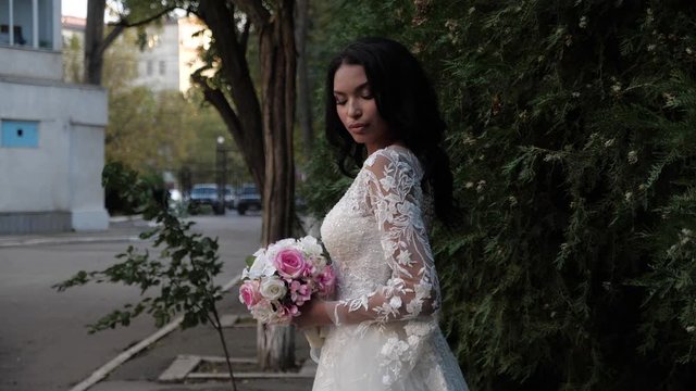 African american lady with languid look in lacy wedding dress holds rose flowers at green bushes against city street slow motion