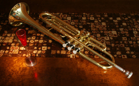 Trompete dourado e ta&ccedil;a vermelha em cima de uma mesa de madeira. Copo de vidro e instrumento musical reflexivo. Mesa de lasca de c&ocirc;co.