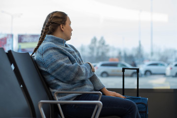 A beautiful girl with hand Luggage sits in the waiting room at the airport and looks out the window sadly. The concept of unwanted departure and sadness after saying goodbye