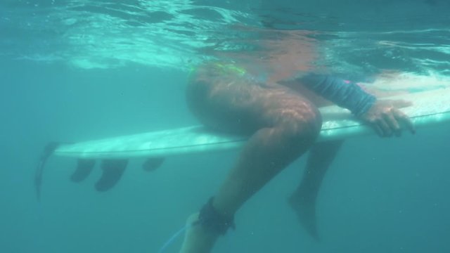 Underwater shot of a surfer girl in a wetsuit seats on a surfboard and waits for a wave in the blue clear pacific ocean water