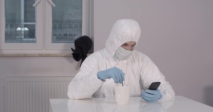 Portrait of young male person stitting behind white dining desk in kitchen in protective suit with a cup of tea and reading news on his phone with a black cat in background during pandemic coronavirus