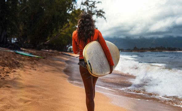 Surfer girl in a wetsuit walks away on the beach, carrying white surfboard