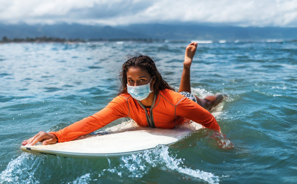 Surfer girl in medical disposable mask and a wetsuit paddling on a surfboard in the ocean.