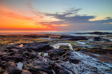 Obraz premium Seascape. Beach with rocks and stones. Low tide. Sun and clouds reflection in water. Slow shutter speed. Soft focus. Tanah Lot beach, Bali, Indonesia