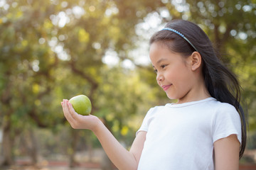 photo happy Little asian girl child standing with big smile. holding green apple in your hand.fresh healthy green bio background with abstract blurred foliage and bright summer sunlight