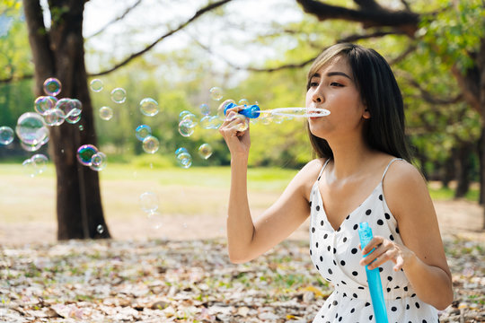 South East Asian ethnic 20s woman blowing air bubbles in the park Young Happy girl wears stylish dress with trees in background. Outdoor summer weekend activity - with copy space