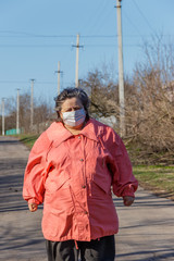 Senior woman stands in a medical mask on her face, which protects against coronavirus and other viruses and diseases on a rural street. Coronavirus pandemic