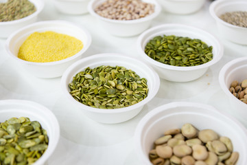 Peeled pumpkin seeds lie in a white plastic plate. Around in the same plates are various seeds, nuts and legumes.