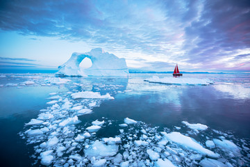 Sail boat with red sails cruising in front of a massive pierced ice berg after sunset. Disko Bay, Greenland. - obrazy, fototapety, plakaty